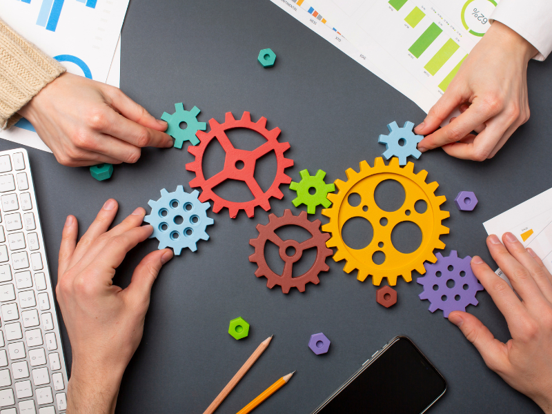 Quatro mãos de pessoas brancas sobre uma mesa , manipulando engrenagens coloridas.