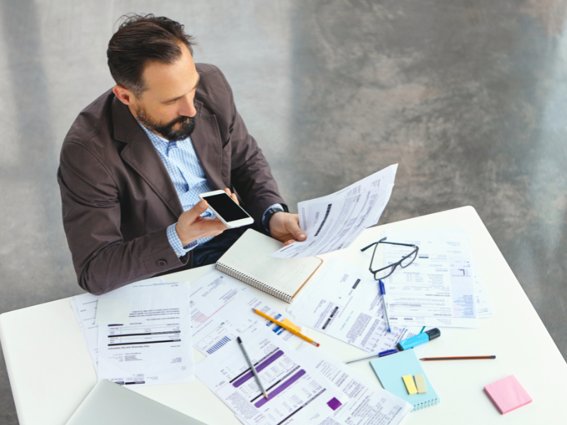 homem branco de paletó, segurando um celular em uma mão e papéis na outra