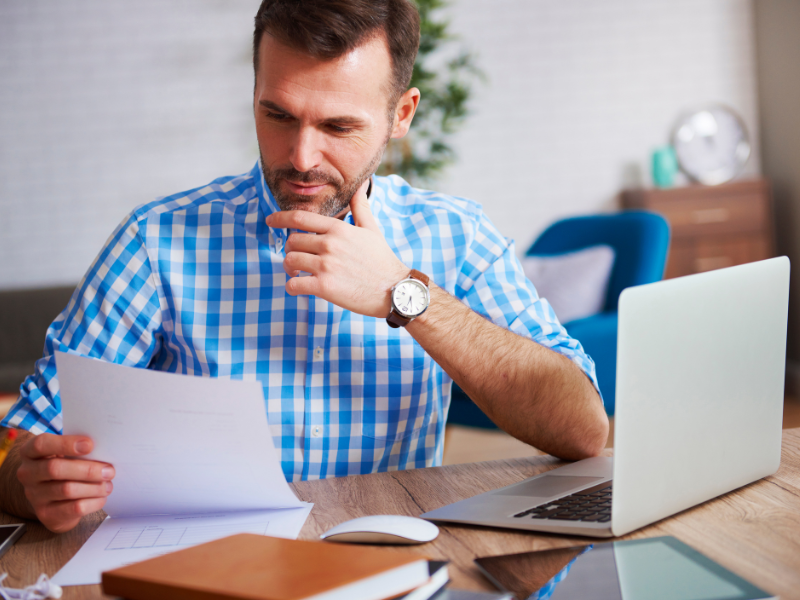 homem branco com relógio no pulso, camisa xadrez azul com branco e segurando papel